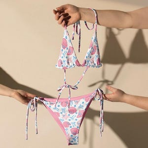 Może przedstawiać: Dwuczęściowy zestaw bikini z białą bazą i wzorem różowych muszli, niebieskich kotwic i innych elementów morskich. Top bikini ma trójkątne miseczki i wiązania, a dół jest dwustronny z różowym wnętrzem.