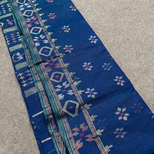 May include: A folded, indigo-blue textile with intricate floral and geometric patterns. The design incorporates pink, white, and gold accents, with vertical stripes and floral motifs. The fabric appears to be lightweight and suitable for various applications.