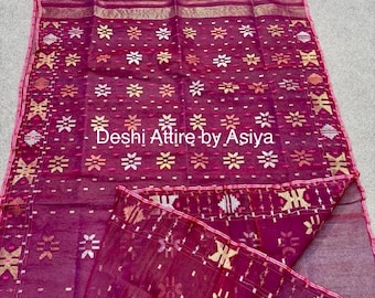 May include: A burgundy sheer saree with a white and gold floral pattern. The saree has a border with a geometric design. The saree is folded and draped on a white surface. "Deshi Attire by Asiya" is written in white text on the saree.