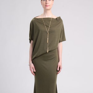 Può includere: Un lungo abito maxi verde oliva con scollo a barca e maniche corte. L'abito è realizzato in un tessuto morbido e fluido e ha una vestibilità rilassata. La modella indossa una collana d'oro con un pendente a nappa.