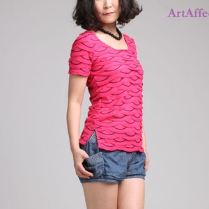 Può includere: Una camicia rosa a maniche corte con un motivo ondulato nero. La camicia ha un collo rotondo e una silhouette leggermente aderente.