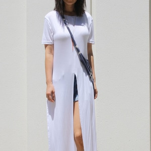 May include: A white long-sleeved t-shirt with a front slit and a long, thin necklace. The shirt is worn over denim shorts and brown sandals.