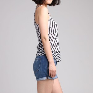 May include: A sleeveless tank top featuring a white base with a bold black geometric line pattern, paired with dark blue denim shorts that have a cuffed hem and visible back pockets. The outfit is shown from a side profile view against a plain background.