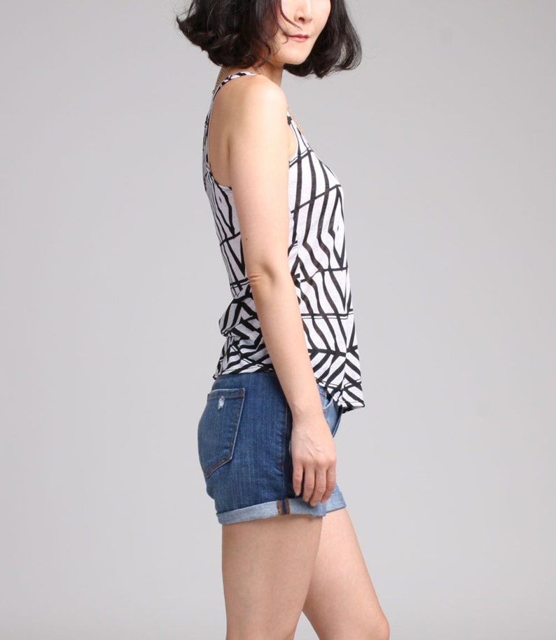 May include: A sleeveless tank top featuring a white base with a bold black geometric line pattern, paired with dark blue denim shorts that have a cuffed hem and visible back pockets. The outfit is shown from a side profile view against a plain background.