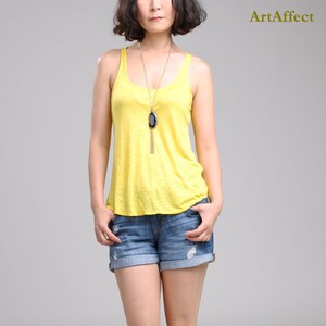 May include: A bright yellow sleeveless tank top with a scoop neckline paired with blue denim shorts featuring distressed details and rolled cuffs. A long gold-tone necklace with a large black agate pendant hangs centered over the yellow top.