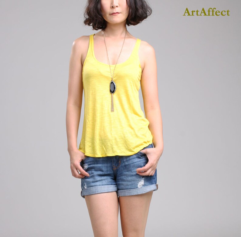 May include: A bright yellow sleeveless tank top with a scoop neckline paired with blue denim shorts featuring distressed details and rolled cuffs. A long gold-tone necklace with a large black agate pendant hangs centered over the yellow top.