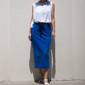 May include: A woman wearing a white sleeveless top and a royal blue pencil skirt. The top has a unique draped design, and the skirt features a black drawstring. White sandals complete the outfit, suitable for casual or semi-formal wear.