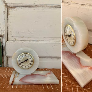 May include: A white marble clock with Roman numeral markings on the face. The clock is on a white marble base with pink and brown streaks. The clock is on a brown and white patterned surface.