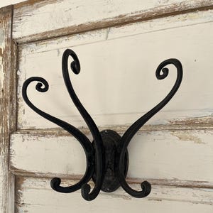 May include: Black metal wall hook with three curved arms. The hook is mounted on a white wooden wall.