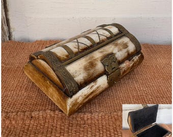 May include: A decorative chest with a hinged lid, featuring a combination of light brown and off-white tones. The chest has metal accents and a clasp closure. The interior is lined with black fabric. The chest is placed on a textured, brown surface.