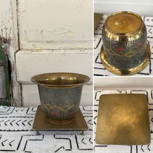 May include: A brass planter with a green and gold floral design. The planter sits on a square brass stand with four legs.