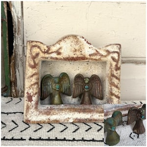 May include: A white ceramic frame with a distressed finish, featuring two small ceramic angels with turquoise accents. The angels are positioned inside the frame, which has a decorative arched top.