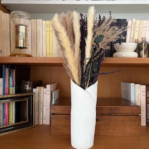 May include: A white cylindrical vase with a textured surface and a folded top edge, containing a dried floral arrangement featuring tall beige pampas grass, dark purple plumes, and dark green eucalyptus stems, standing upright on a wooden shelf.