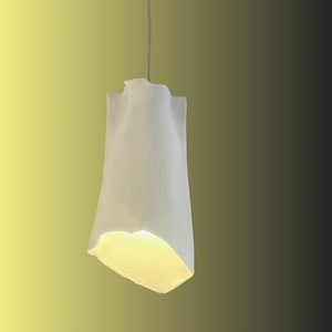 Peut inclure: Un luminaire suspendu en tissu blanc avec une forme froissée et organique. La lumière est allumée et émet une lueur jaune chaude.