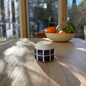 May include: A small, white candle holder with a geometric design of dark blue and brown squares. The candle holder sits on a light-colored wooden table. A bowl of oranges and greenery is in the background.