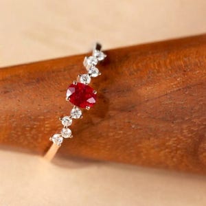 May include: A delicate ring featuring a central oval-cut red gemstone flanked by a series of small, round, clear white stones set along a thin, polished gold band that follows a slightly curved, asymmetrical design.