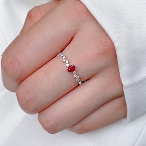 May include: A delicate silver-toned ring featuring a central oval-cut red gemstone flanked by a series of small, clear round-cut stones arranged in a linear pattern along the thin metal band, creating a refined and balanced appearance.