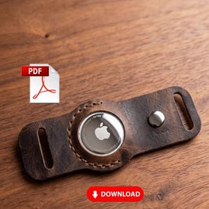 Könnte beinhalten: Ein brauner Leder-AirTag-Halter mit silbernem Knopfverschluss. Der AirTag ist in der Mitte sichtbar, und der Halter hat an jedem Ende eine Schlaufe. Der Hintergrund ist eine Holzoberfläche. Das Wort "DOWNLOAD" ist sichtbar.