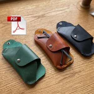 Puede incluir: Tres estuches de gafas de cuero en verde, marrón y negro, cada uno con cierre de botón a presión. El estuche marrón contiene un par de gafas. Los estuches están sobre una superficie de madera. Un icono PDF está en la esquina superior izquierda.