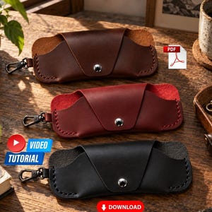 Op de afbeelding: Drie lederen brillenkokers in bruin, rood en zwart. Elke koker heeft een driehoekige flap met een zilverkleurige metalen drukknoopsluiting, zichtbare stiksels langs de randen en een zwarte metalen draaibare karabijnhaak aan één zijkant.
