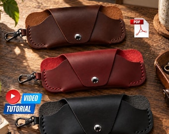Puede incluir: Tres estuches de cuero para gafas en marrón, rojo y negro. Cada estuche presenta una solapa triangular con un cierre de botón a presión de metal plateado, costuras visibles en los bordes y un mosquetón giratorio de metal negro en un lado.