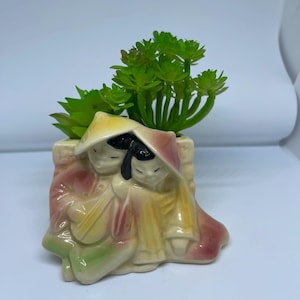 Puede incluir: Una maceta de cerámica con una planta suculenta verde, que representa dos figuras con vestimenta tradicional. La maceta tiene un fondo beige con detalles en amarillo, rosa y verde. Las figuras llevan sombreros y túnicas.