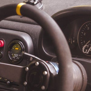 Peut inclure: Un volant noir de rechange avec un rep&egrave;re central jaune sur le dessus, plac&eacute; devant le tableau de bord d'une voiture. Le combin&eacute; d'instruments comporte un compte-tours avec le texte GAS GHOST et une jauge AEM air-carburant circulaire.