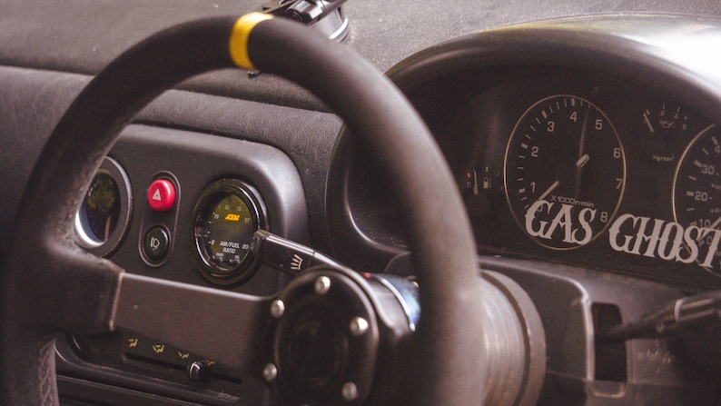 Peut inclure: Un volant noir de rechange avec un rep&egrave;re central jaune sur le dessus, plac&eacute; devant le tableau de bord d'une voiture. Le combin&eacute; d'instruments comporte un compte-tours avec le texte GAS GHOST et une jauge AEM air-carburant circulaire.