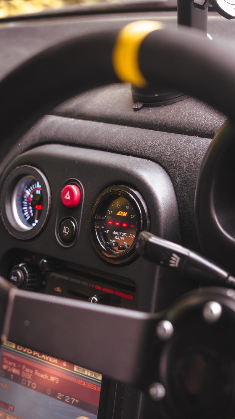 Peut inclure: Un int&eacute;rieur de tableau de bord de voiture avec un volant noir dot&eacute; d'un rep&egrave;re sup&eacute;rieur jaune, une jauge de rapport air/carburant AEM circulaire avec affichage num&eacute;rique rouge, un bouton de feux de d&eacute;tresse rouge et un lecteur DVD.
