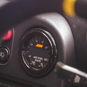 Peut inclure: Un manom&egrave;tre de rapport air/carburant AEM circulaire mont&eacute; sur un tableau de bord noir. L'instrument pr&eacute;sente un affichage num&eacute;rique avec des graduations de 8 &agrave; 20 et le logo AEM en orange. Deux petits boutons MODE et SEL sont situ&eacute;s en dessous.