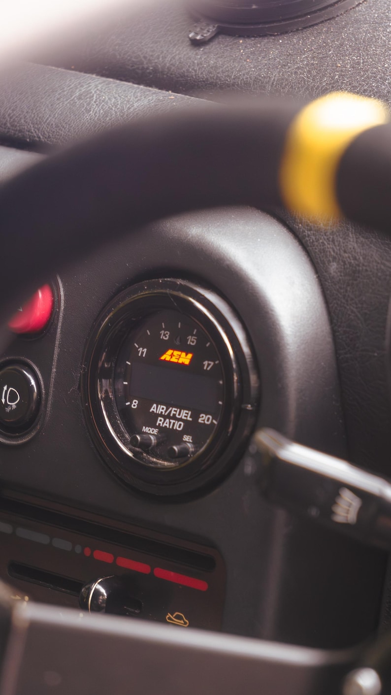 Peut inclure: Un manom&egrave;tre de rapport air/carburant AEM circulaire mont&eacute; sur un tableau de bord noir. L'instrument pr&eacute;sente un affichage num&eacute;rique avec des graduations de 8 &agrave; 20 et le logo AEM en orange. Deux petits boutons MODE et SEL sont situ&eacute;s en dessous.