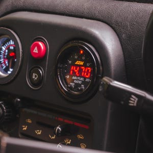 Peut inclure: Un manom&egrave;tre de tableau de bord automobile noir affichant une lecture num&eacute;rique rouge de 14,70 pour le rapport air/carburant. Le manom&egrave;tre circulaire de marque AEM est mont&eacute; dans une console en plastique sombre pr&egrave;s d'un bouton de feux de d&eacute;tresse.