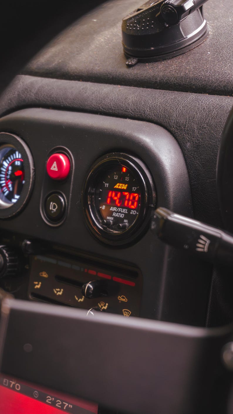 Peut inclure: Un manom&egrave;tre de tableau de bord automobile noir affichant une lecture num&eacute;rique rouge de 14,70 pour le rapport air/carburant. Le manom&egrave;tre circulaire de marque AEM est mont&eacute; dans une console en plastique sombre pr&egrave;s d'un bouton de feux de d&eacute;tresse.