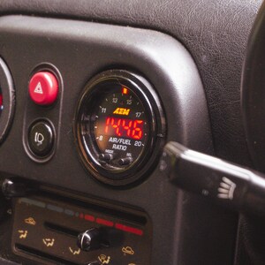 Peut inclure: Une console centrale de tableau de bord automobile noire comprenant un manom&egrave;tre num&eacute;rique AEM air/carburant affichant 14.46 en rouge, un bouton de feux de d&eacute;tresse rouge, un interrupteur de phares, un bouton A/C et des curseurs de climatisation.