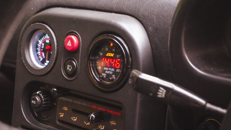 Peut inclure: Une console centrale de tableau de bord automobile noire comprenant un manom&egrave;tre num&eacute;rique AEM air/carburant affichant 14.46 en rouge, un bouton de feux de d&eacute;tresse rouge, un interrupteur de phares, un bouton A/C et des curseurs de climatisation.