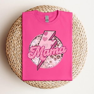 以下が含まれることがあります： 「Mama」の文字がバブルフォントで描かれた明るいピンク色のTシャツ。ピンク色の稲妻とハートのアクセント、ヒョウ柄の円がデザインされています。Tシャツは編み込まれた表面に折りたたまれています。