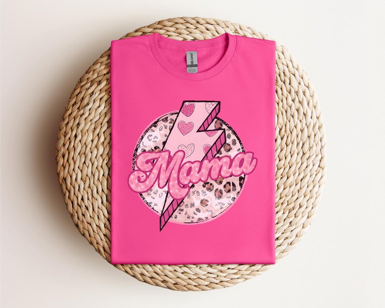 以下が含まれることがあります： 「Mama」の文字がバブルフォントで描かれた明るいピンク色のTシャツ。ピンク色の稲妻とハートのアクセント、ヒョウ柄の円がデザインされています。Tシャツは編み込まれた表面に折りたたまれています。