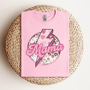 以下が含まれることがあります： 明るいピンク色のTシャツ。デザインは、より濃いピンクで縁取られたピンク色の「ママ」という言葉が特徴です。ハートとヒョウ柄のディテールが施された稲妻が、円形のフレーム内に配置されています。