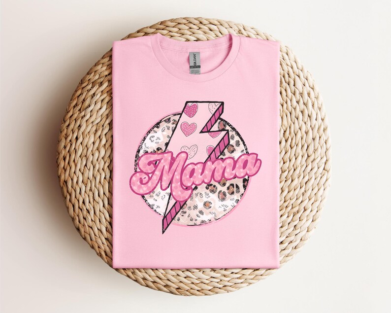 以下が含まれることがあります： 明るいピンク色のTシャツ。デザインは、より濃いピンクで縁取られたピンク色の「ママ」という言葉が特徴です。ハートとヒョウ柄のディテールが施された稲妻が、円形のフレーム内に配置されています。