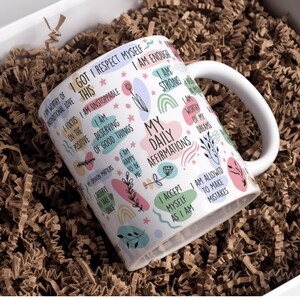 Op de afbeelding: Witte keramische mok met de tekst "My Daily Affirmations" en diverse positieve affirmaties in pastelkleuren. De mok heeft een wit oor en staat in een doos met bruine papieren vulling.