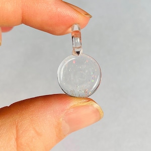 May include: A small, circular, clear resin pendant featuring embedded iridescent glitter particles that reflect light in various colors. The pendant has a transparent loop at the top for attachment, showcasing a smooth, polished, and translucent finish.