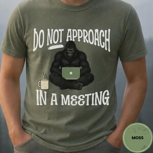 May include: An olive green short-sleeve t-shirt featuring a graphic of a black gorilla sitting and using a light green laptop. Next to the gorilla is a white coffee mug. The text reads DO NOT APPROACH IN A MEETING in white capital letters.