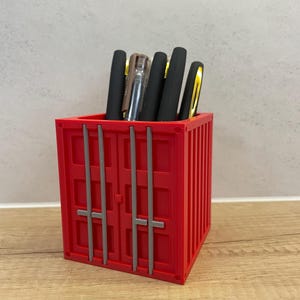 Può includere: Un organizer da scrivania rosso a forma di container che contiene diverse penne. L'organizer ha una forma rettangolare con linee verticali e barre grigie sul davanti. Le penne sono nere e dorate, con una penna trasparente visibile.