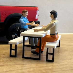Op de afbeelding: Twee plastic actiefiguren zitten tegenover elkaar aan een witte picknicktafel met een zwart metalen frame. Eén figuur draagt een blauw overhemd en een grijze broek, de andere een beige overhemd en een oranje broek. Een rode speelgoedtruck staat erachter.