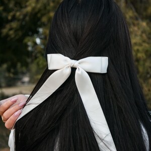 May include: A white satin bow hair accessory is tied into long, dark hair. The bow is centered and has long, flowing tails. The hair is parted in the middle and cascades down the back. The background is blurred with green and brown tones.