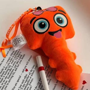 Op de afbeelding: Oranje pluche sleutelvormig speelgoed met grote blauwe ogen, een roze tong en een lus om op te hangen. Een witte pen met rode accenten en een bedrukt papier met tekst staan ook op de afbeelding.