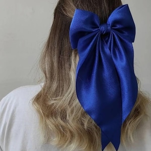 Puede incluir: Un gran lazo de satén azul atado en una forma clásica, usado en el cabello de una persona con cabello largo, ondulado y castaño claro.