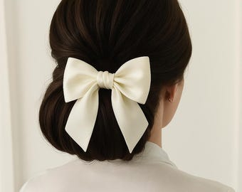 May include: A large, ivory-colored satin bow hair accessory. The bow is centered on the back of a person's dark brown hair, styled in a low bun. The bow's fabric has a subtle sheen, and the overall aesthetic is elegant and classic.