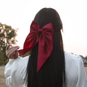 Puede incluir: Un gran lazo de satén burdeos está sujeto en un cabello largo y negro. El lazo es el punto focal, con su diseño de gran tamaño y su color intenso. La persona lleva una blusa blanca y el fondo está borroso.