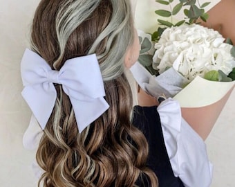May include: A white bow with a large, soft, and fluffy texture is tied in a girl's brown hair. The girl is wearing a black dress and is holding a bouquet of white hydrangeas.